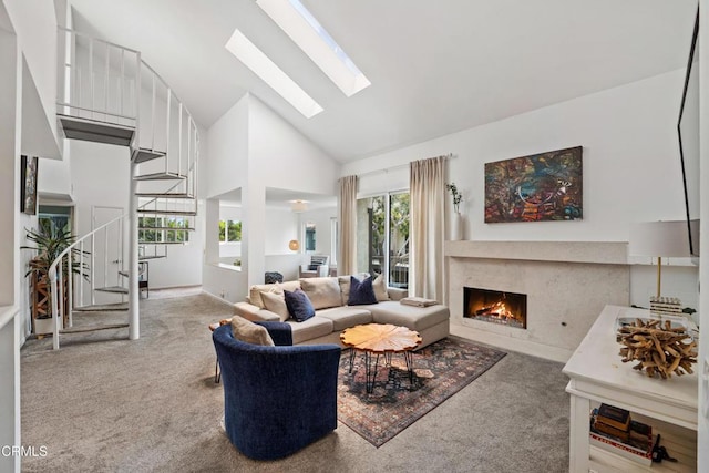 carpeted living area with a lit fireplace, stairs, a skylight, and high vaulted ceiling