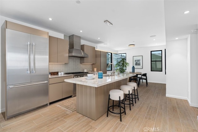 kitchen featuring wall chimney range hood, built in fridge, gas stove, light wood finished floors, and a breakfast bar
