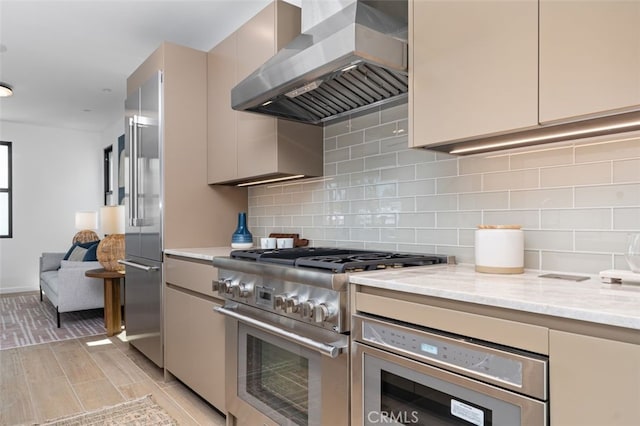 kitchen with wall chimney range hood, premium appliances, light stone counters, and tasteful backsplash