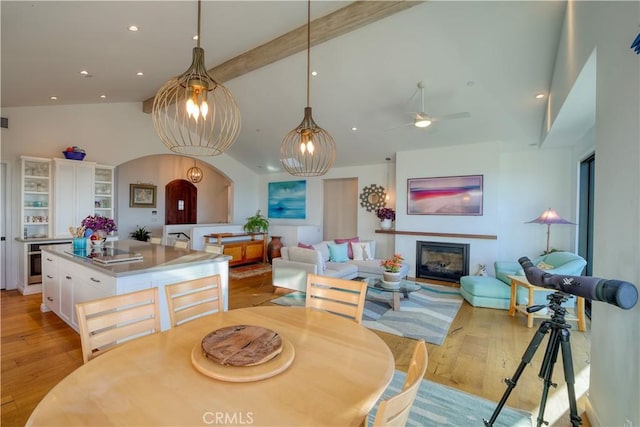 dining room featuring ceiling fan, arched walkways, light wood-type flooring, lofted ceiling with beams, and a glass covered fireplace
