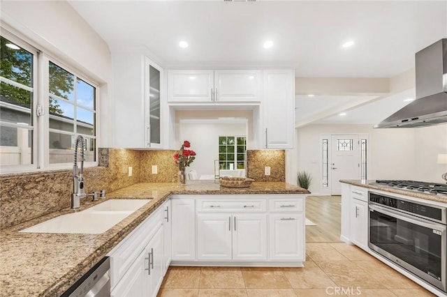 kitchen featuring light stone countertops, wall chimney exhaust hood, a sink, backsplash, and stainless steel appliances