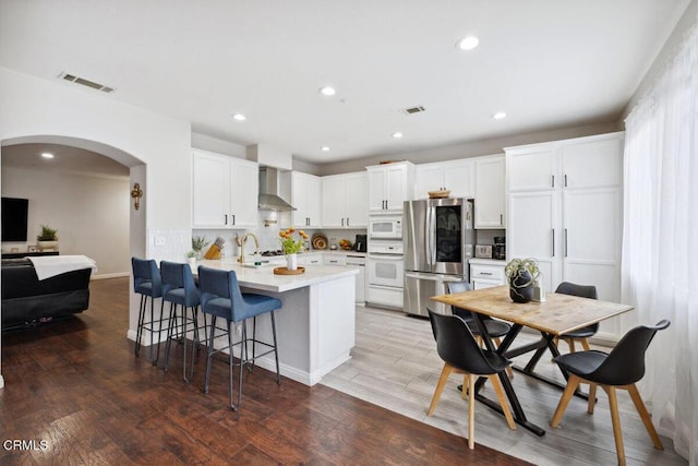 kitchen featuring wall chimney range hood, white appliances, arched walkways, a peninsula, and visible vents