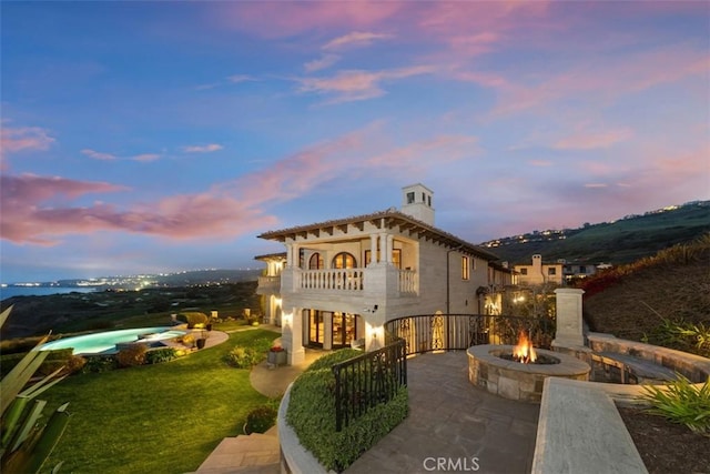 back of property at dusk with an outdoor fire pit, a chimney, a balcony, a lawn, and a patio