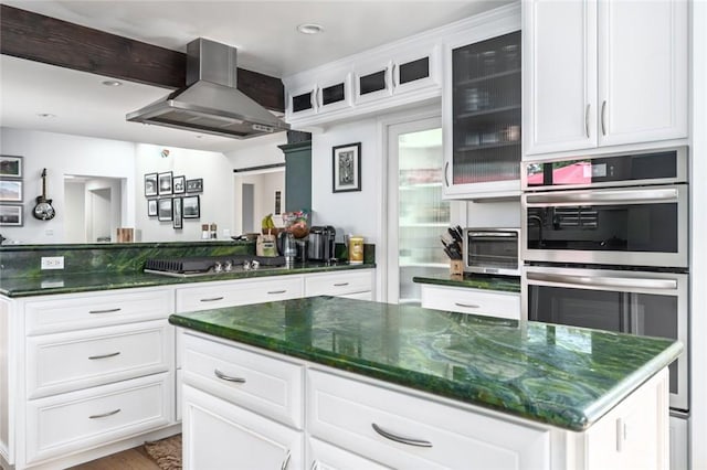 kitchen featuring stainless steel double oven, wall chimney exhaust hood, dark stone counters, and white cabinets