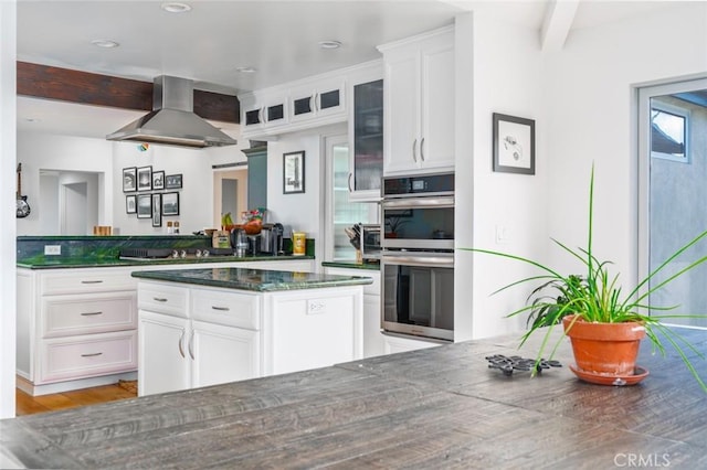 kitchen with double oven, light wood-style floors, wall chimney exhaust hood, white cabinetry, and dark stone countertops