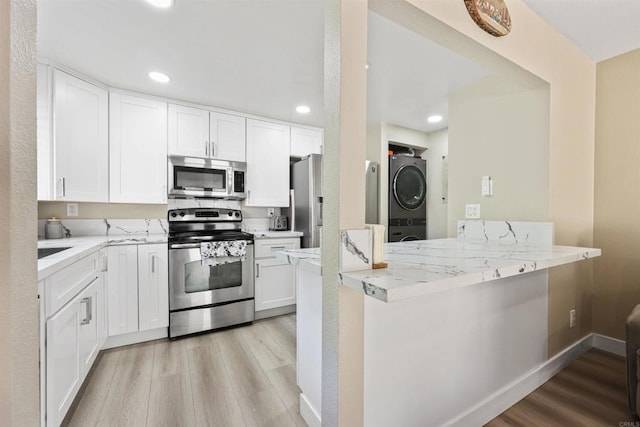 kitchen with a peninsula, stacked washer / dryer, light wood-type flooring, white cabinetry, and appliances with stainless steel finishes