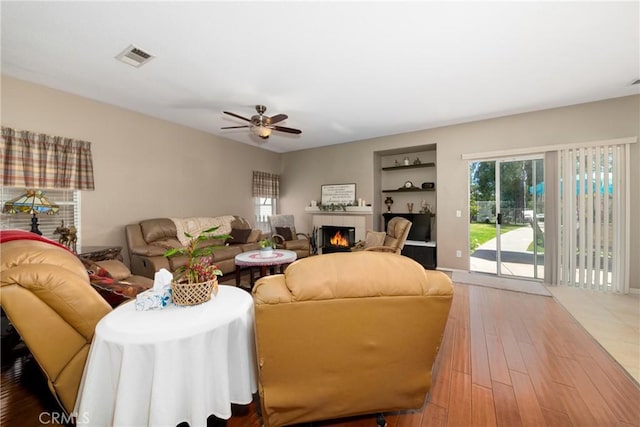 living area featuring ceiling fan, visible vents, built in features, wood finished floors, and a lit fireplace