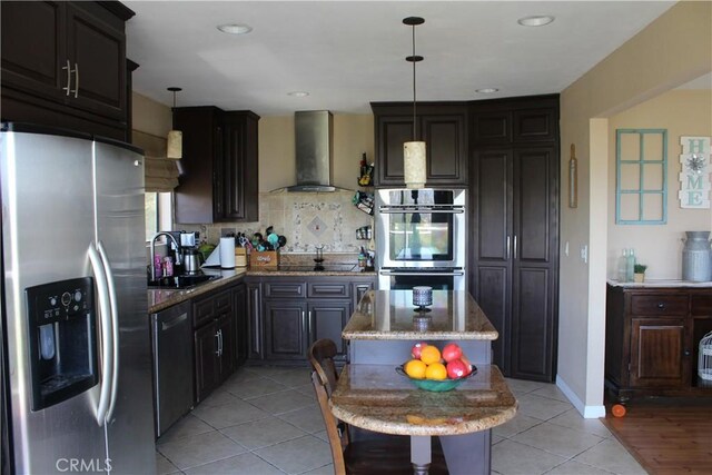kitchen with appliances with stainless steel finishes, hanging light fixtures, a sink, light stone counters, and wall chimney exhaust hood