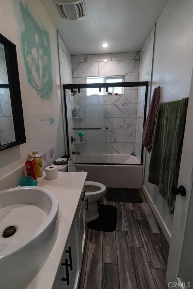 bathroom featuring visible vents, vanity, toilet, wood finish floors, and combined bath / shower with glass door