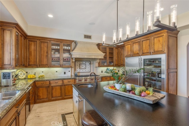 kitchen with brown cabinetry, stainless steel built in fridge, hanging light fixtures, glass insert cabinets, and custom range hood