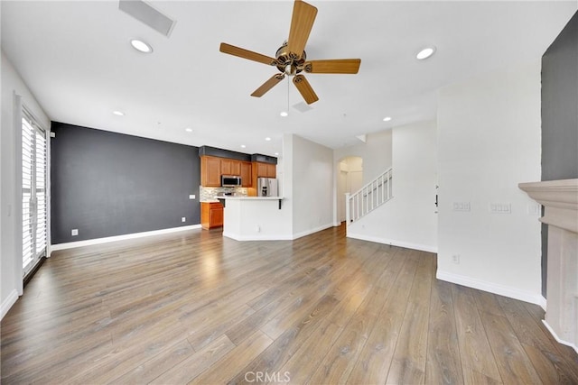 unfurnished living room with light wood-style floors, arched walkways, stairs, a ceiling fan, and baseboards