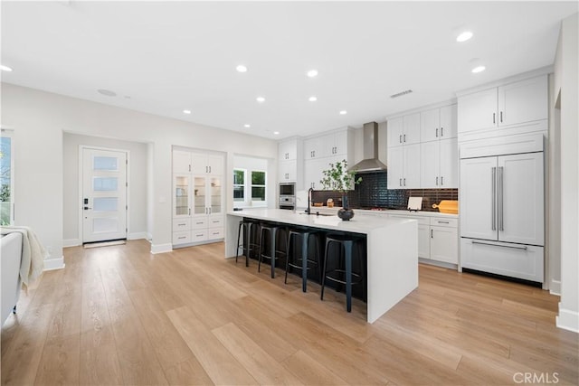 kitchen with white cabinets, wall chimney range hood, built in appliances, a breakfast bar area, and a large island with sink