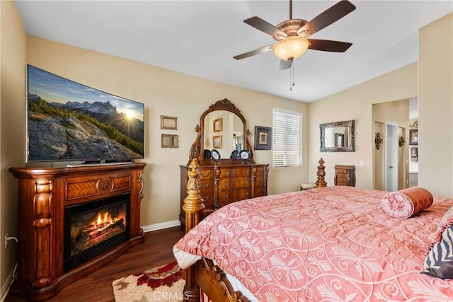 bedroom featuring a glass covered fireplace, baseboards, ceiling fan, dark wood finished floors, and vaulted ceiling