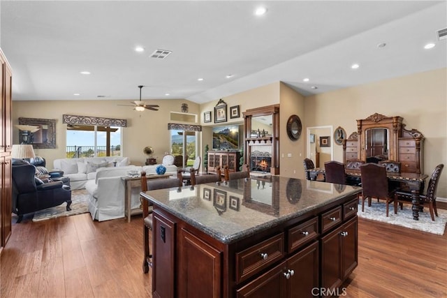 kitchen with dark wood-style flooring, a ceiling fan, a lit fireplace, vaulted ceiling, and visible vents