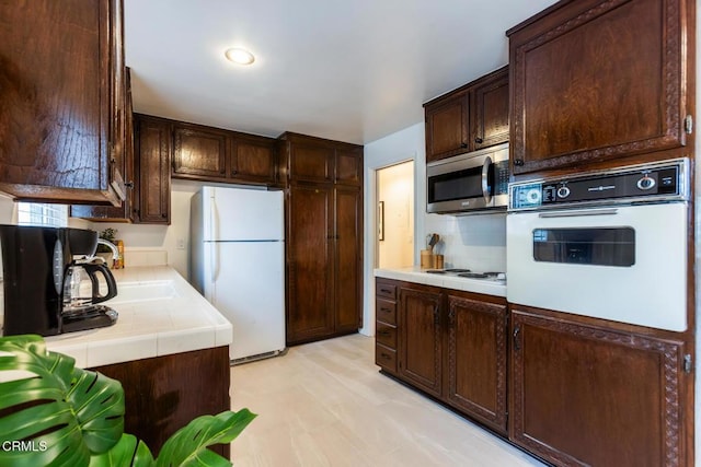 kitchen featuring decorative backsplash, dark brown cabinetry, tile countertops, a sink, and white appliances
