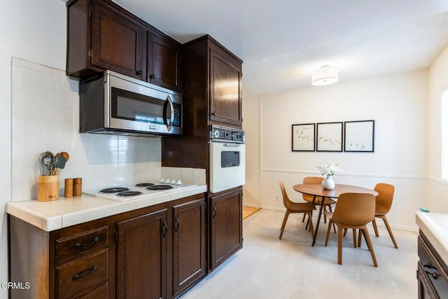kitchen with dark brown cabinetry, tile countertops, tasteful backsplash, baseboards, and white appliances