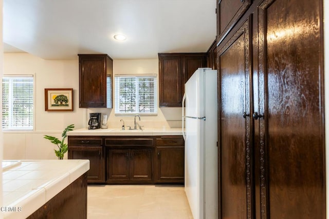 kitchen featuring freestanding refrigerator, dark brown cabinets, tile counters, a wainscoted wall, and a sink