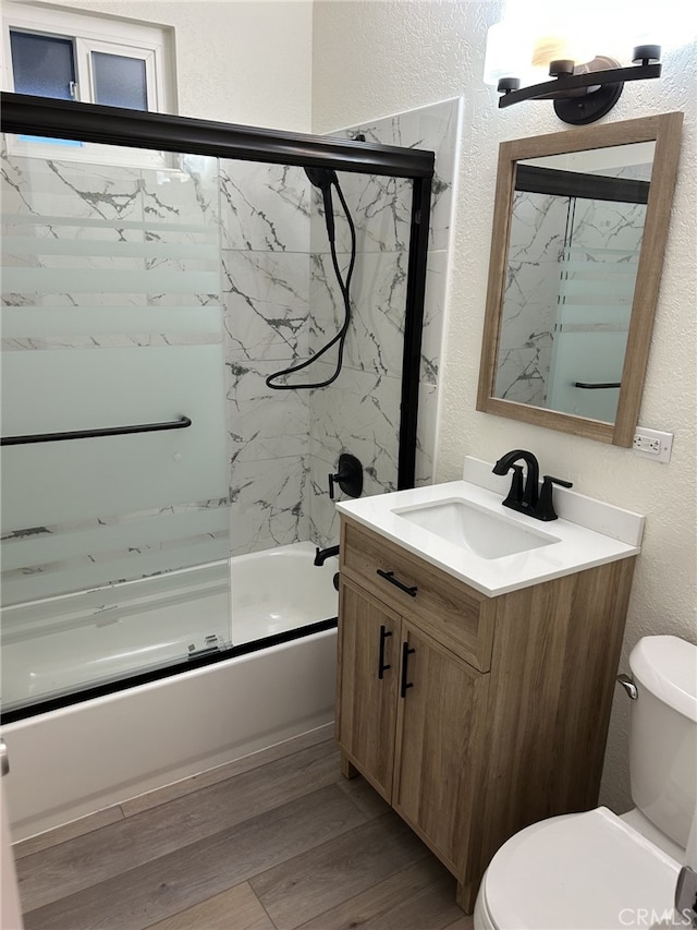 bathroom featuring a textured wall, toilet, wood finished floors, shower / bath combination with glass door, and vanity