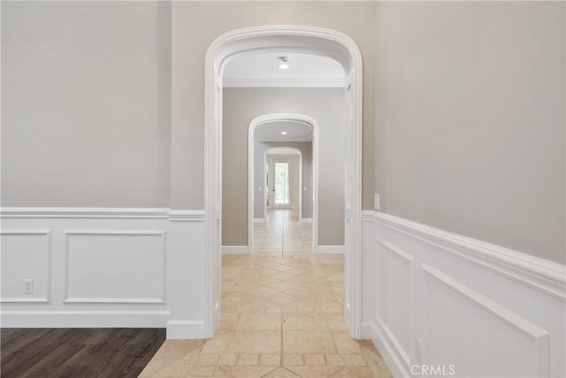 corridor with crown molding, arched walkways, a wainscoted wall, and a decorative wall