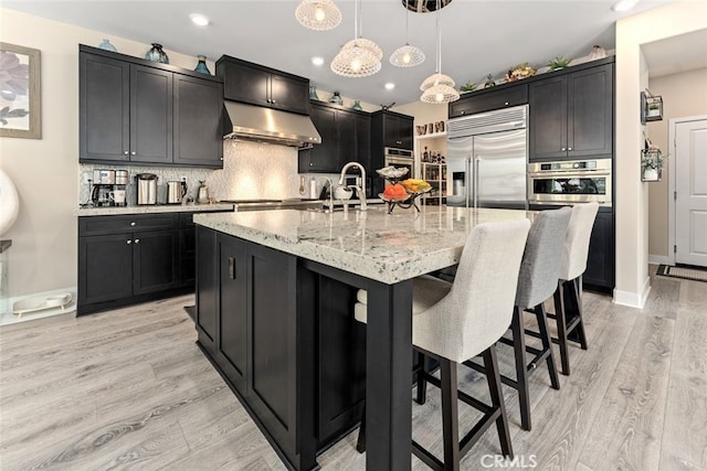 kitchen with light stone counters, light wood-style floors, stainless steel appliances, decorative backsplash, and pendant lighting