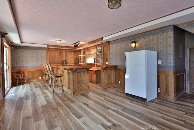 kitchen featuring freestanding refrigerator, wainscoting, wallpapered walls, and brown cabinetry