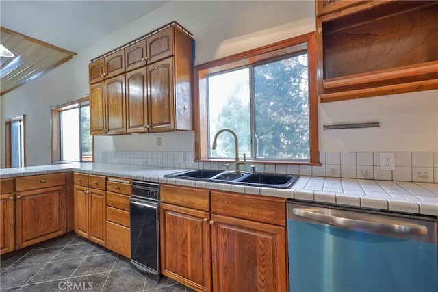 kitchen with a sink, tile counters, brown cabinetry, dark tile patterned floors, and dishwashing machine