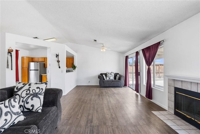 living room featuring a ceiling fan, a textured ceiling, vaulted ceiling, a tile fireplace, and wood finished floors