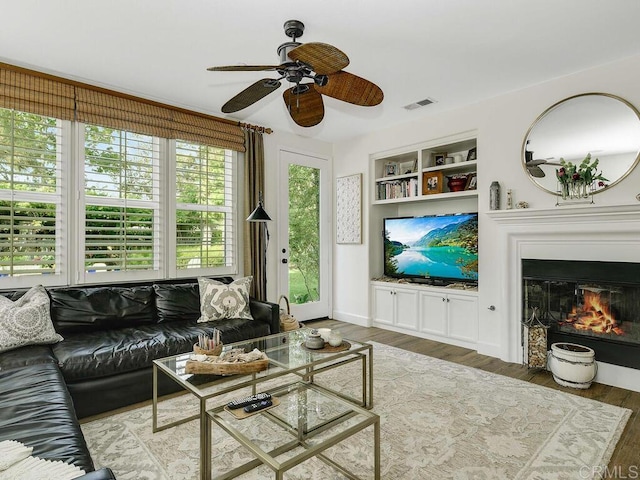 living room with a ceiling fan, visible vents, built in features, wood finished floors, and a glass covered fireplace