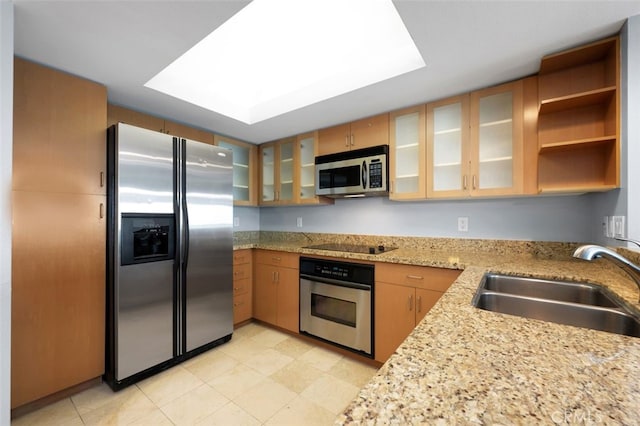 kitchen with brown cabinetry, appliances with stainless steel finishes, light stone countertops, a sink, and glass insert cabinets