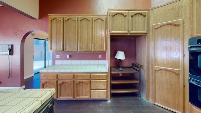 kitchen featuring dobule oven black, tile countertops, arched walkways, dark wood finished floors, and a textured wall