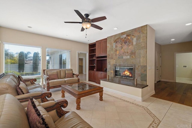 living room with baseboards, ceiling fan, built in shelves, a tiled fireplace, and light tile patterned floors