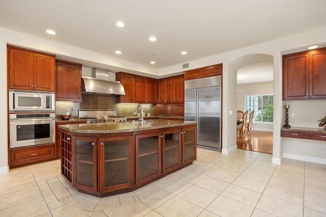 kitchen with built in appliances, light tile patterned flooring, backsplash, and wall chimney exhaust hood