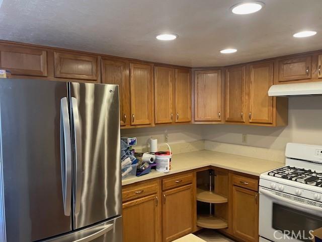 kitchen with light countertops, brown cabinetry, freestanding refrigerator, under cabinet range hood, and white range with gas cooktop