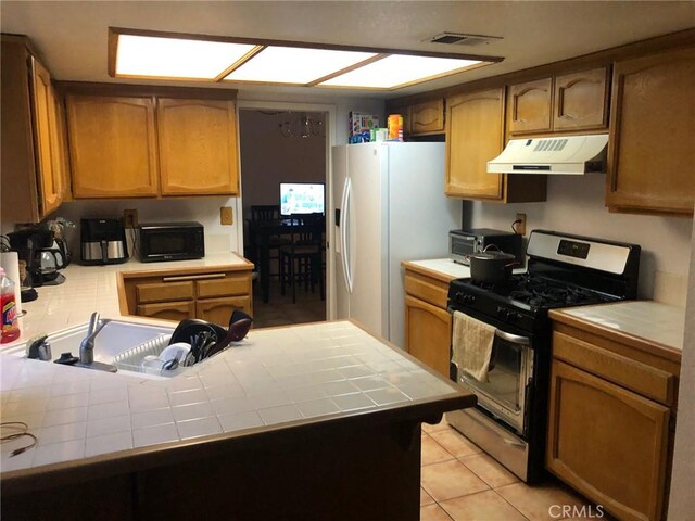 6483 N Walnut Ave, San Bernardino, CA 92407 - kitchen featuring stainless steel gas range, tile counters, under cabinet range hood, a peninsula, and white fridge with ice dispenser