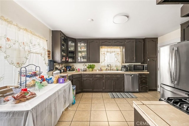 kitchen with dark brown cabinets, appliances with stainless steel finishes, a sink, light tile patterned floors, and glass insert cabinets
