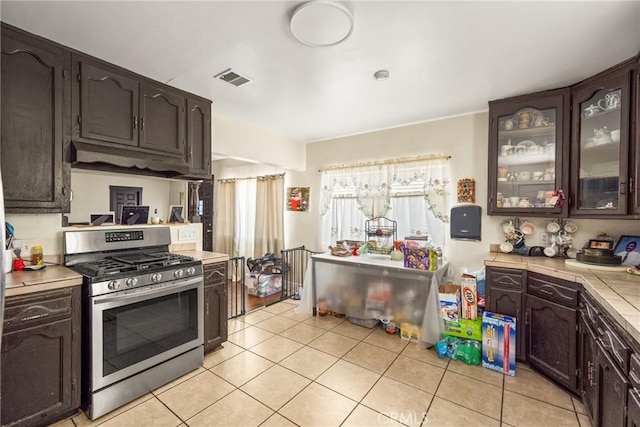 kitchen with light tile patterned flooring, dark brown cabinetry, gas stove, visible vents, and glass insert cabinets