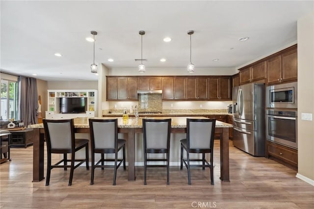 kitchen featuring stainless steel appliances, under cabinet range hood, light stone counters, decorative light fixtures, and light wood-style floors