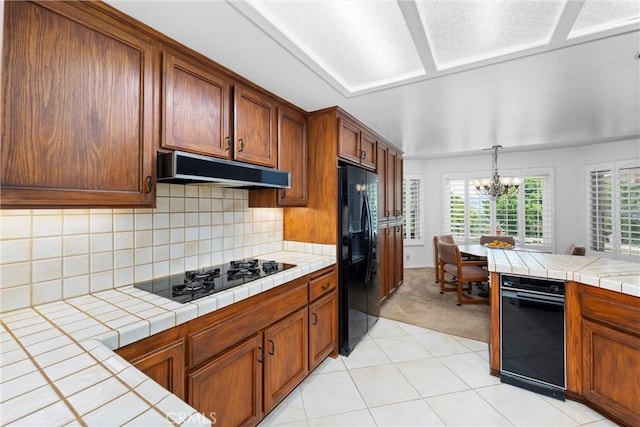 kitchen with an inviting chandelier, tile counters, under cabinet range hood, brown cabinets, and black appliances