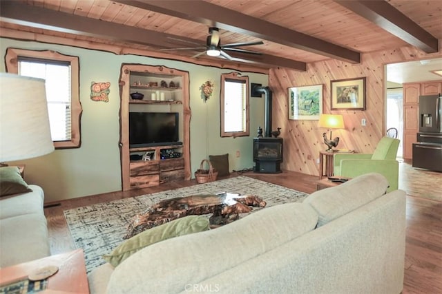living room featuring a ceiling fan, wood walls, wood finished floors, a wood stove, and beam ceiling