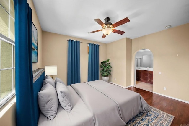 bedroom featuring a ceiling fan, arched walkways, wood finished floors, baseboards, and connected bathroom