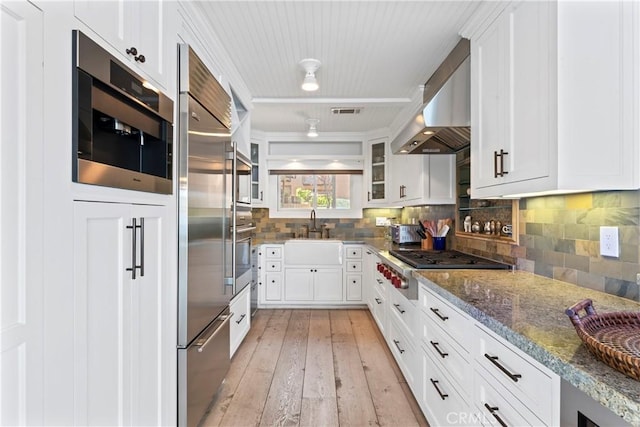 kitchen with a sink, wall chimney exhaust hood, dark stone countertops, white cabinetry, and visible vents