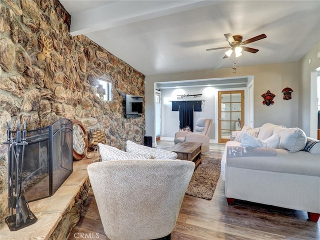 living room with beam ceiling, wood finished floors, a ceiling fan, and a stone fireplace