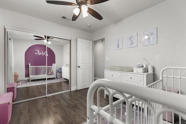 bedroom with a closet, a nursery area, dark wood finished floors, ceiling fan, and visible vents