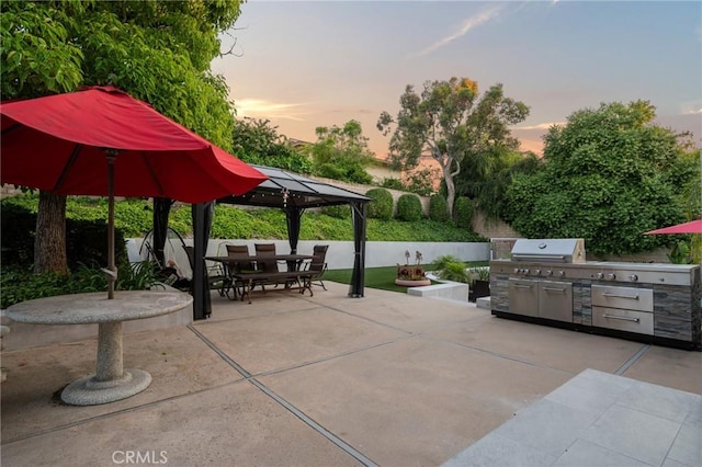 patio terrace at dusk featuring outdoor dining area, a gazebo, grilling area, and exterior kitchen