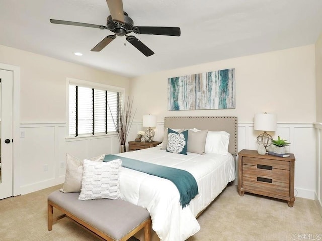bedroom with light carpet, a decorative wall, ceiling fan, and wainscoting