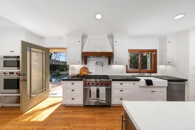 kitchen featuring a sink, white cabinetry, premium range hood, light wood-style floors, and appliances with stainless steel finishes