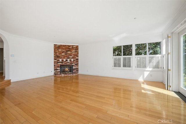 unfurnished living room featuring arched walkways, light wood finished floors, a brick fireplace, ornamental molding, and baseboards