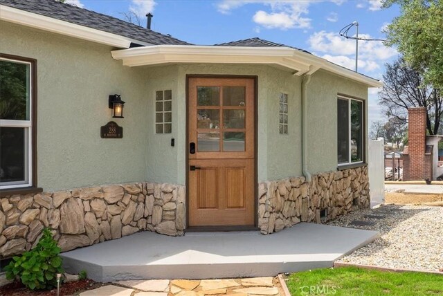doorway to property featuring stone siding, stucco siding, and a shingled roof