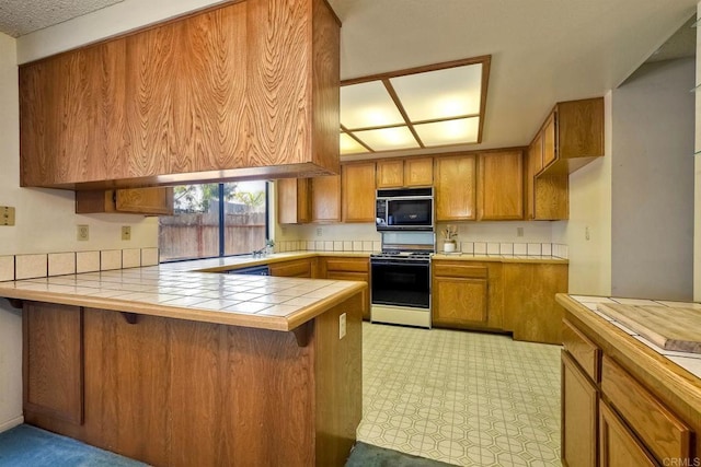 kitchen featuring brown cabinetry, gas range, black microwave, a peninsula, and tile counters