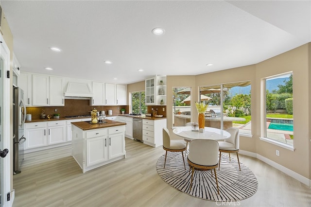 kitchen with appliances with stainless steel finishes, custom range hood, a kitchen island, backsplash, and light wood-type flooring